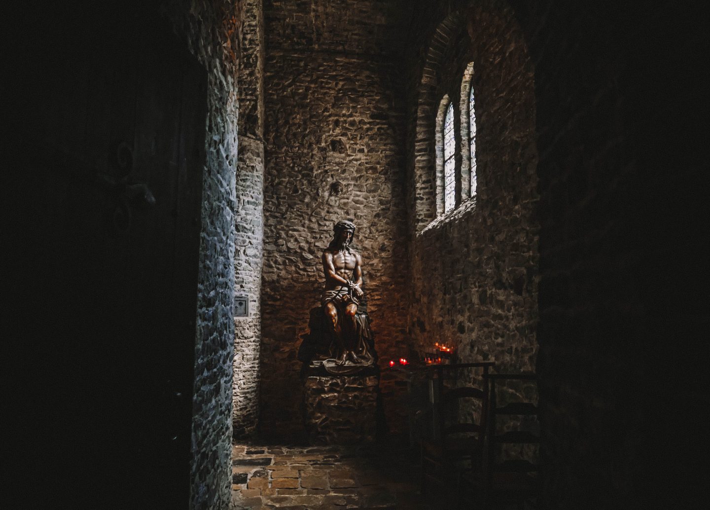 A dimly lit stone chapel in Bruges, featuring a wooden statue of Christ seated with a crown of thorns, illuminated by soft natural light from a nearby arched window. The rugged stone walls and scattered lit candles create a solemn and contemplative atmosphere, emphasizing the spiritual and historic ambiance of the sacred space.