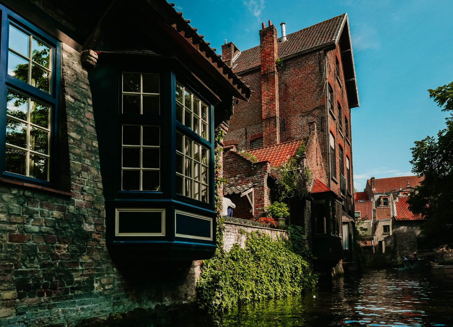 A charming canal-side scene in Bruges, featuring historic brick buildings with large windows and ivy-covered walls. A bay window painted in dark blue extends over the water, reflecting the sunlight. The red-tiled roofs and greenery add to the picturesque, storybook-like atmosphere of this medieval Belgian town.