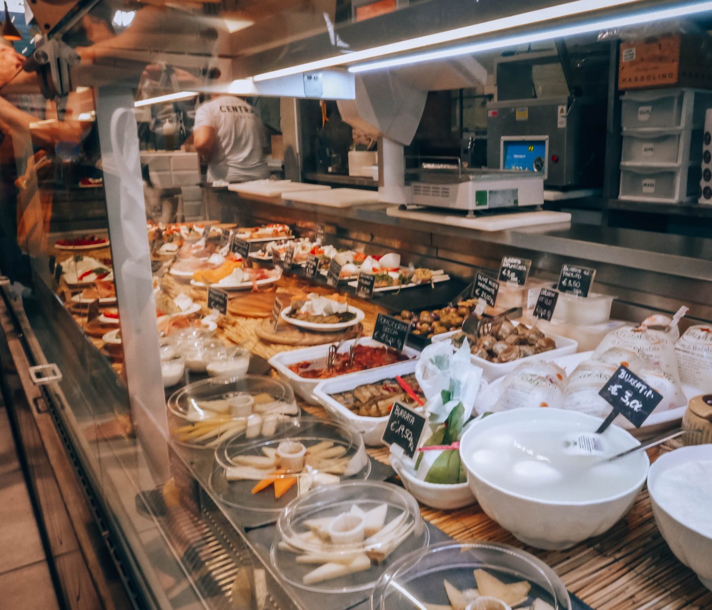 This image showcases a glass display case at Mercato Centrale in Florence, filled with a variety of fresh Italian cheeses, appetisers, and gourmet delicacies. The selection includes burrata, aged cheeses, marinated vegetables, bruschetta, and small plates of antipasti, each labeled with handwritten price tags. The reflection on the glass adds to the bustling market ambiance, with a worker in the background preparing more food. A true paradise for food lovers!
