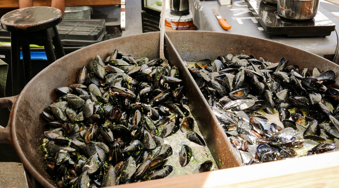 This image captures a large cast-iron pan filled with freshly cooked mussels, served at Odprta Kuhna, the vibrant open-air food market in Ljubljana, Slovenia. The mussels are glistening in a flavourful broth, garnished with fresh herbs, and divided into sections in the pan. In the background, a rustic food stall setup with a portable stove, knives, and cooking equipment enhances the market’s street-food atmosphere. The dish showcases the fresh, high-quality seafood offerings available at this bustling culinary event.