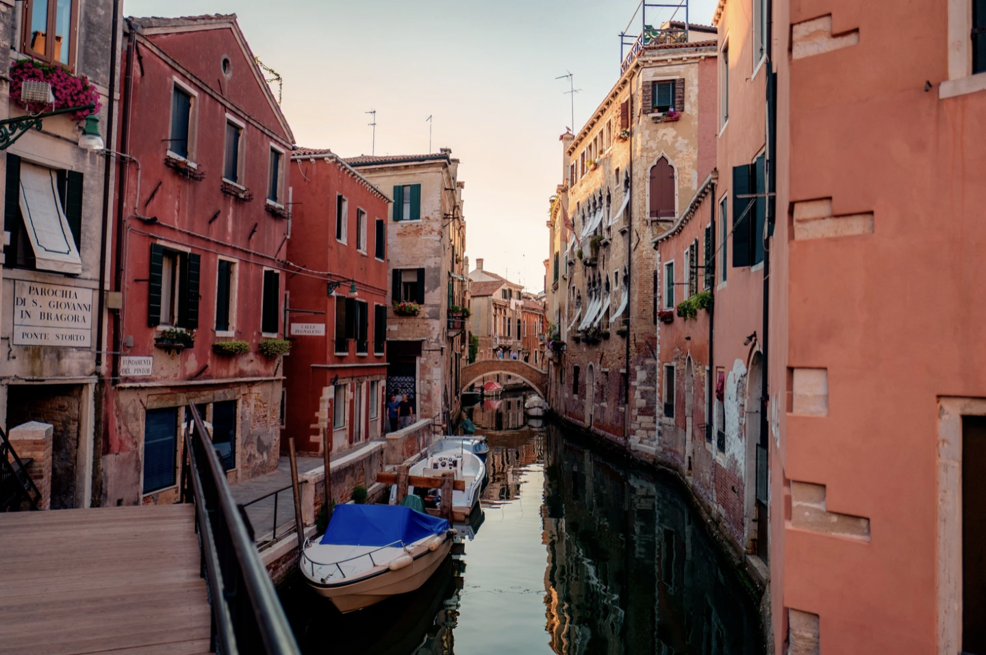 Charming canal scene in Venice, Italy at sunset, featuring small boats moored along the waterway lined with colourful, weathered buildings. A narrow footbridge arches over the canal in the distance, with flower boxes and laundry adding local character to the windows and balconies.