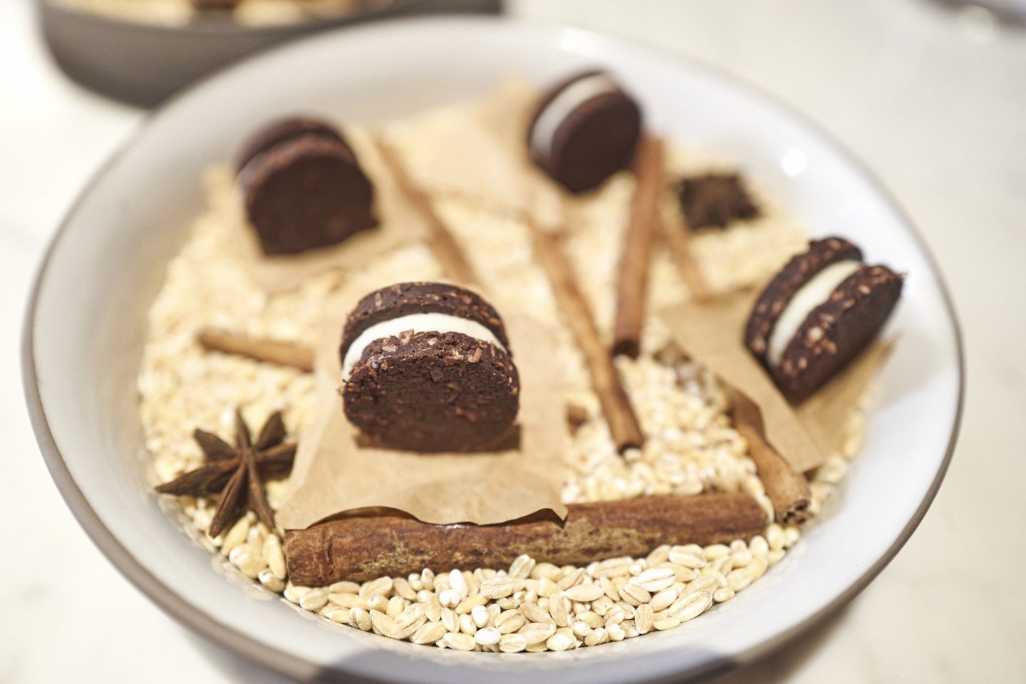 A beautifully plated dessert at Michelin starred Essencia Restaurant in Budapest, featuring small sandwich cookies with a creamy white filling. The cookies are arranged on brown parchment paper over a bed of barley grains, cinnamon sticks, and star anise, creating a rustic and aromatic presentation.