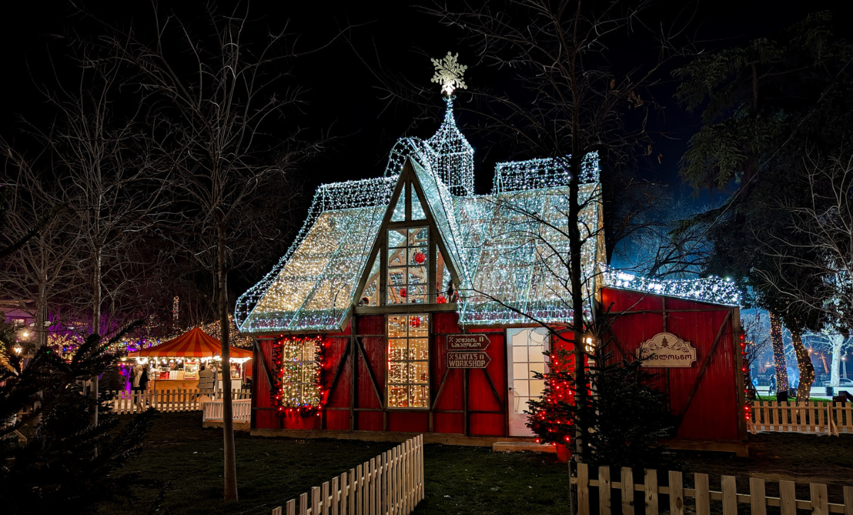 A festive Christmas cabin in Georgia, adorned with twinkling lights shaped like a house labeled 'Santa's Workshop,' enclosed by a white picket fence. Nearby, a red and yellow tent and illuminated trees enhance the magical holiday ambiance at night.