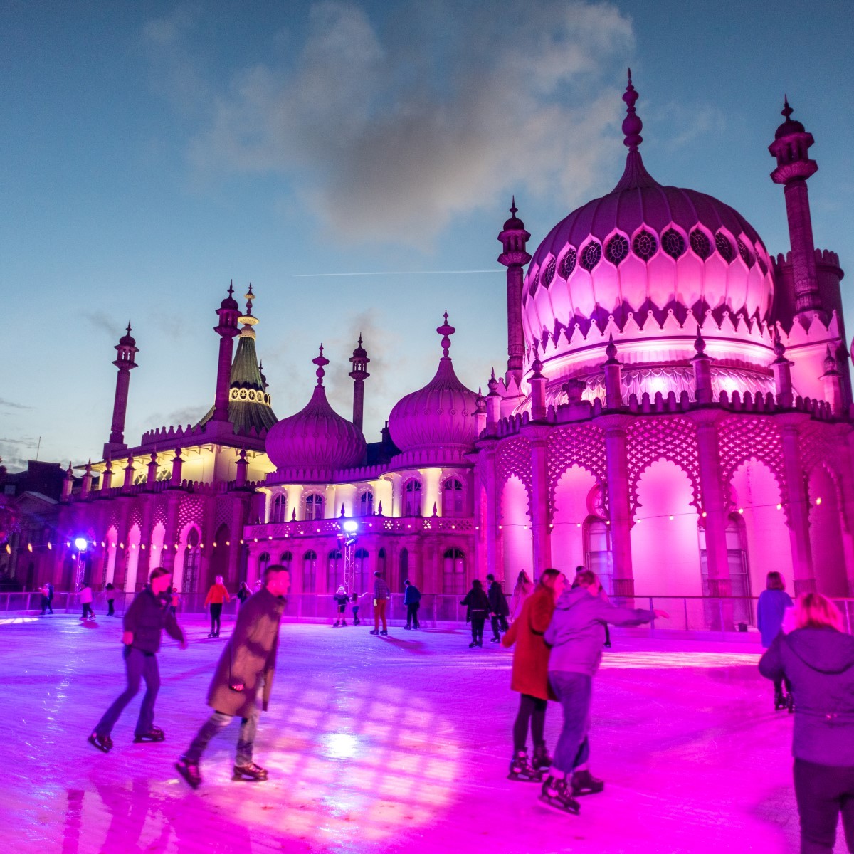 People ice skating at the Brighton Pavilion ice rink in the UK, with the iconic Indian-style domes and arches of the pavilion illuminated in pink and purple lights against the twilight sky.