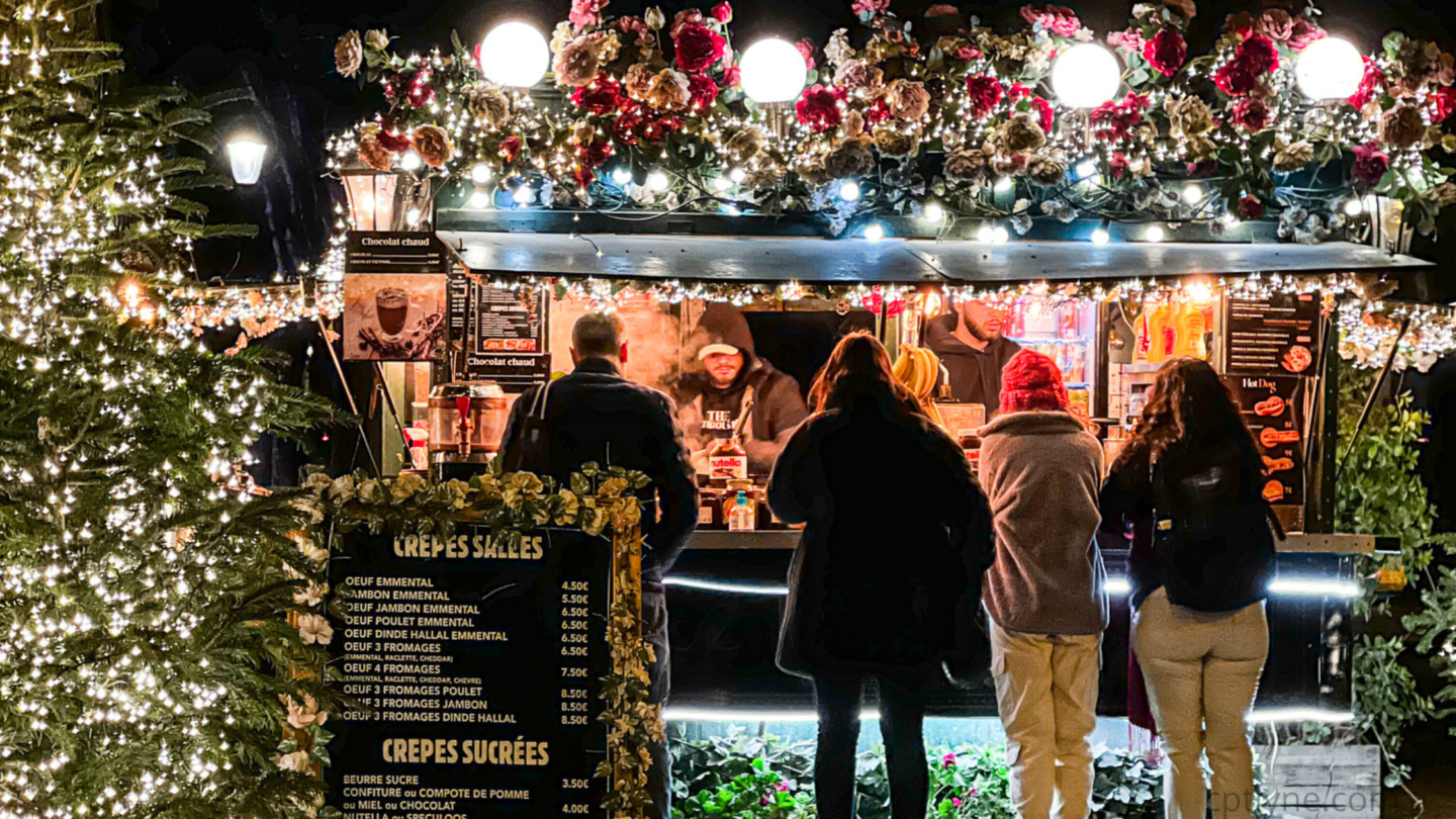 People gather at a French Christmas market food stall decorated with flowers and lights, with a menu listing various crepes, and a lit Christmas tree nearby enhancing the holiday atmosphere.