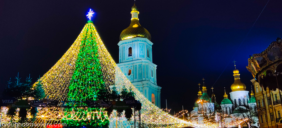 A towering Christmas tree adorned with green and gold lights stands in front of the historic St. Sophia's Cathedral in Kyiv, Ukraine, with its iconic golden domes glowing under the night sky, creating a festive and spiritual holiday scene.