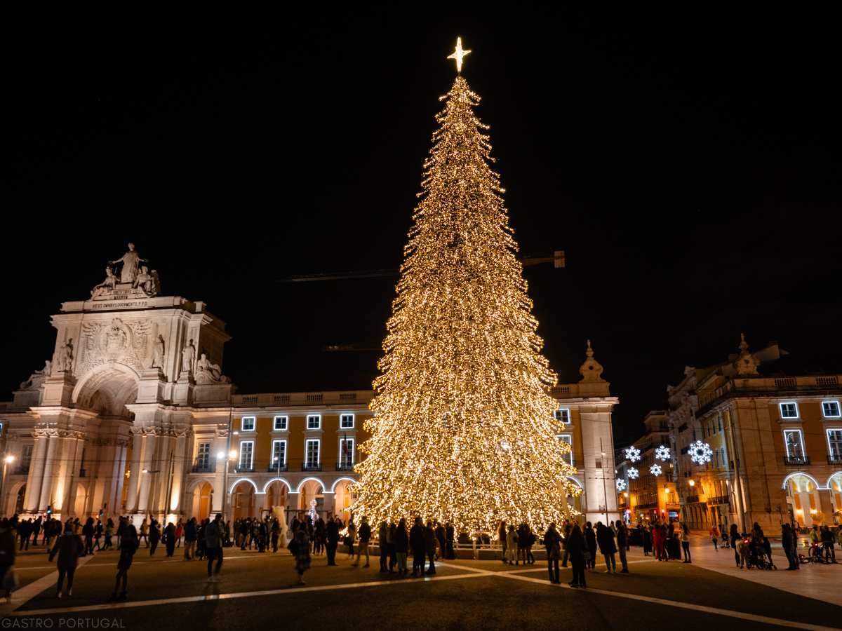 A towering Christmas tree illuminated with golden lights and topped with a glowing star stands in Praça do Comércio, Lisbon, Portugal, with the ornate Arco da Rua Augusta and surrounding historic buildings creating a festive backdrop.