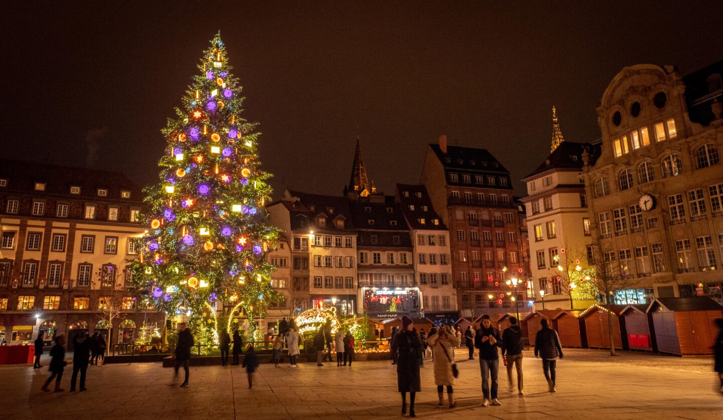 A large, illuminated Christmas tree in a square in Strasbourg, France, surrounded by historic buildings and people enjoying the festive atmosphere at night.
