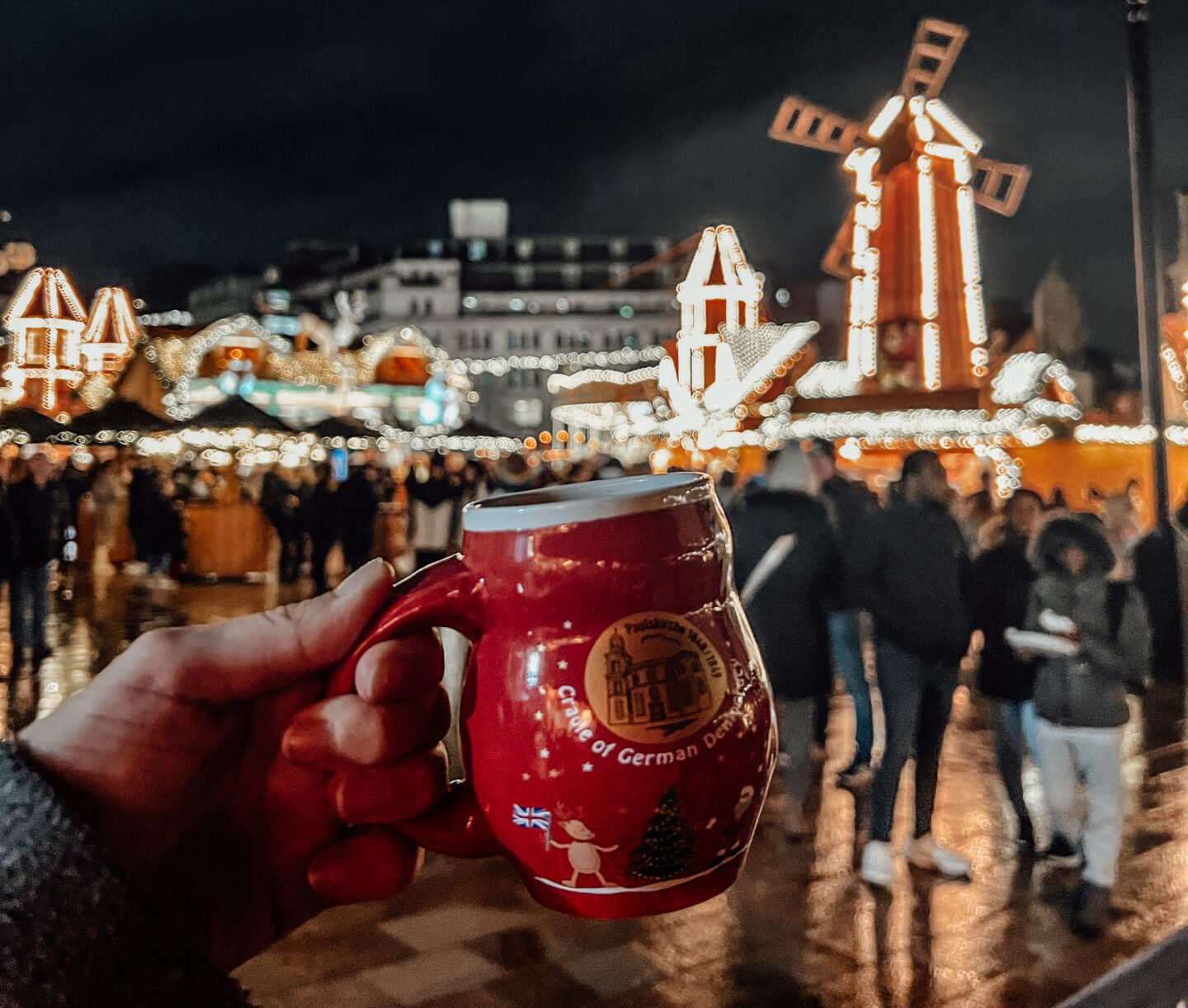 A hand holding a festive red mug with "German Christmas Market" branding, framed by the market’s glowing lights and wooden stalls in the background.