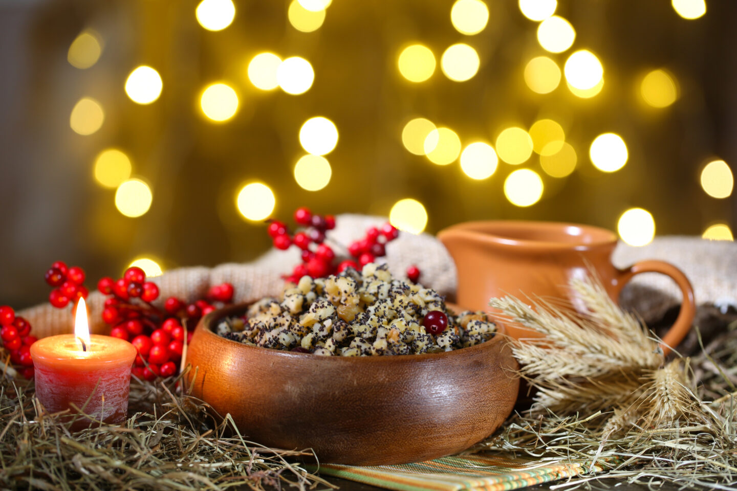 A bowl of traditional Ukrainian kutya, a sweet grain pudding made with wheat, poppy seeds, nuts, and honey, sits surrounded by a lit candle, red berries, and wheat stalks against a background of soft, golden holiday lights.