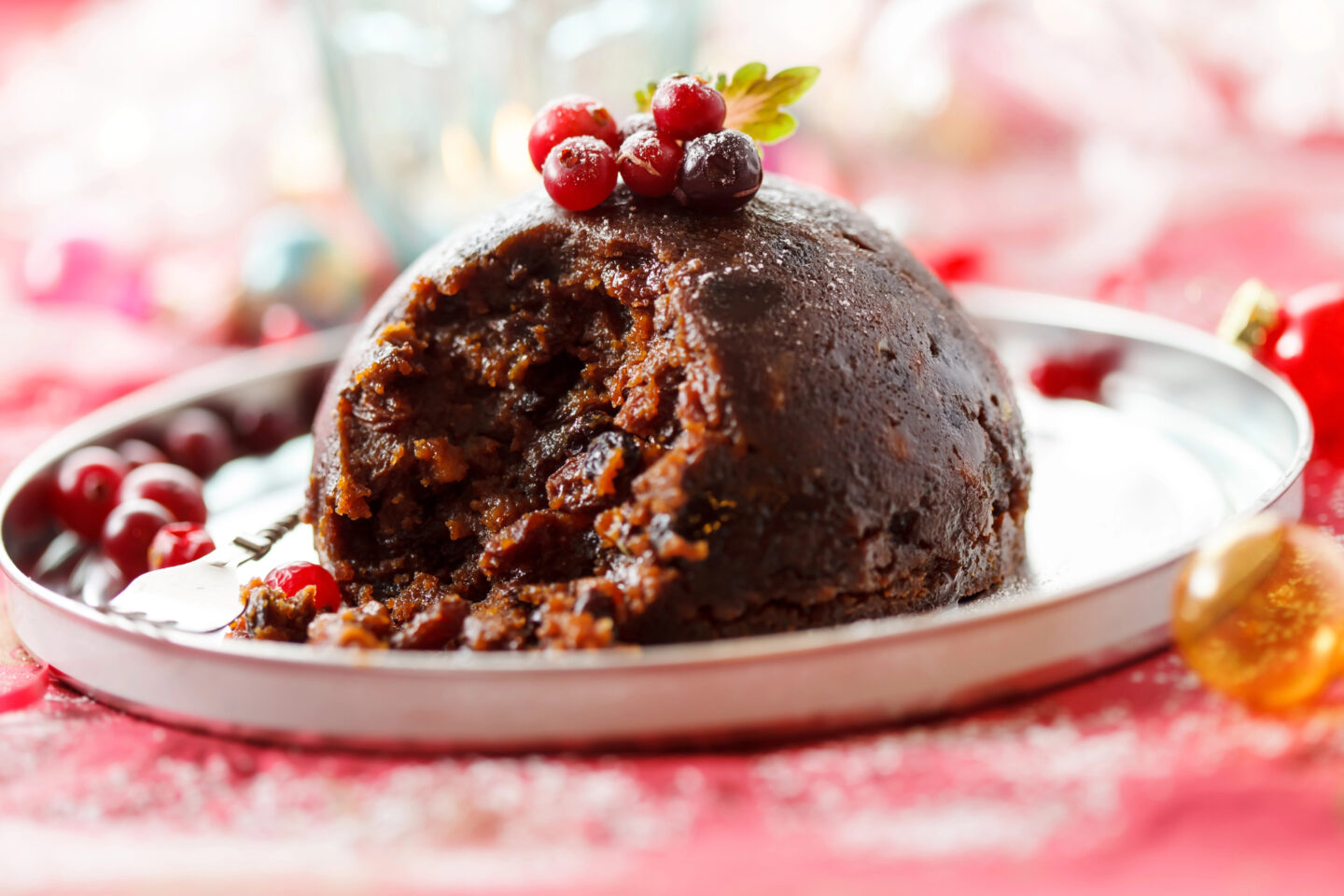 A traditional Christmas pudding served on a silver plate, garnished with red berries on top, showcasing its rich, dark texture with a bite taken out, capturing a classic British holiday dessert.