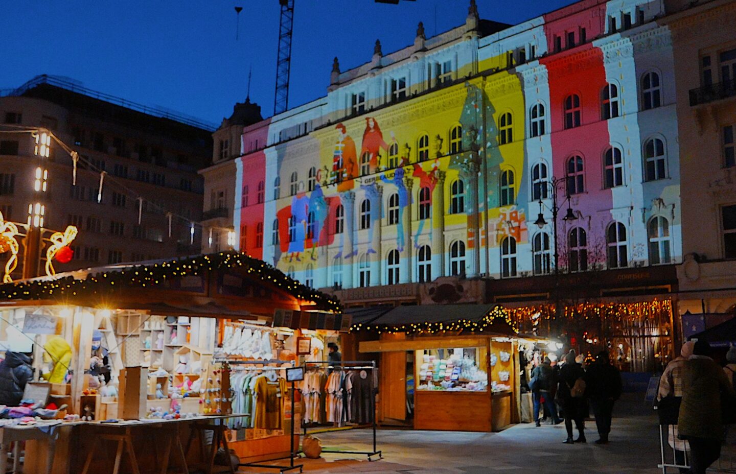 A Christmas market in Budapest, Hungary, with festive wooden stalls illuminated by string lights, selling holiday items. A building in the background is decorated with colourful holiday-themed projections, adding a vibrant touch to the nighttime scene.