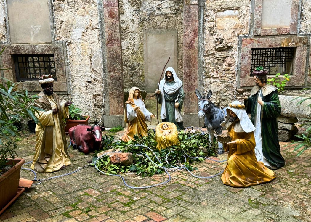 A traditional outdoor Nativity scene with statues of Mary, Joseph, the baby Jesus, and the Three Wise Men, along with a donkey and cow, set against an old stone wall with greenery on the ground, capturing the essence of a rustic Christmas celebration.