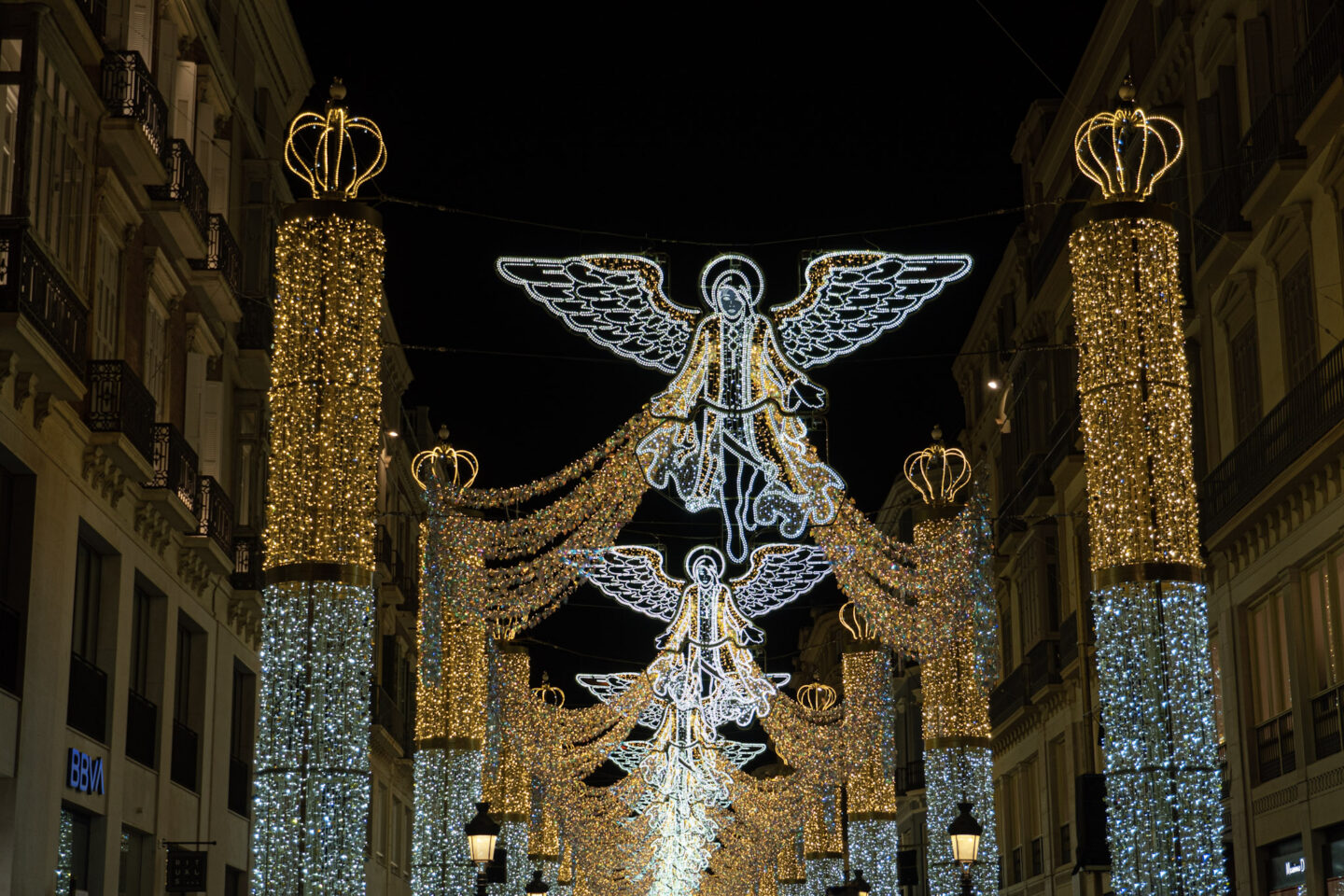 Christmas lights in Malaga illuminate a street, featuring glowing angel figures suspended above and large decorative pillars wrapped in golden and silver lights topped with crown-shaped ornaments. The angel lights, with intricate detailing, hover in a line down the street, creating a festive and majestic holiday atmosphere against the night sky.