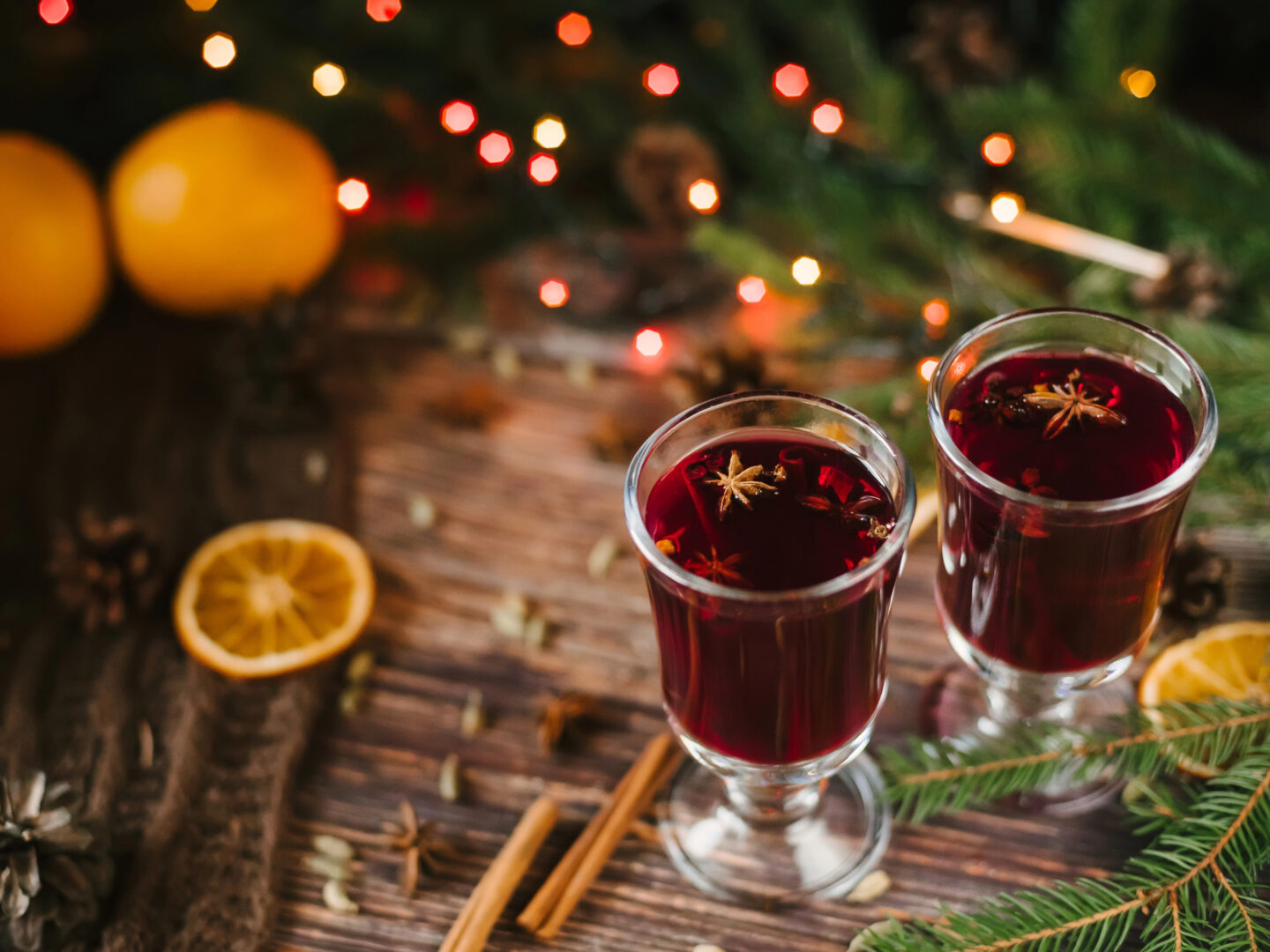 Two glasses of mulled wine (Glühwein) garnished with star anise sit on a wooden table surrounded by festive decorations, including pine branches, cinnamon sticks, and orange slices, with a warm glow from blurred Christmas lights in the background.
