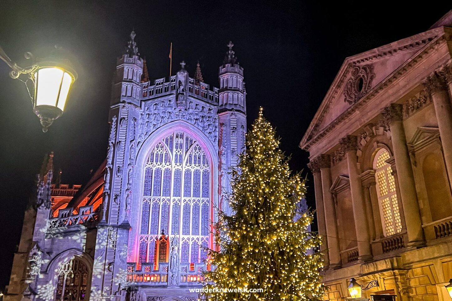 Bath Abbey illuminated at night with festive lighting, standing next to a large Christmas tree adorned with golden lights. A traditional street lamp glows in the foreground, adding to the cozy holiday atmosphere at the Bath Christmas Market.