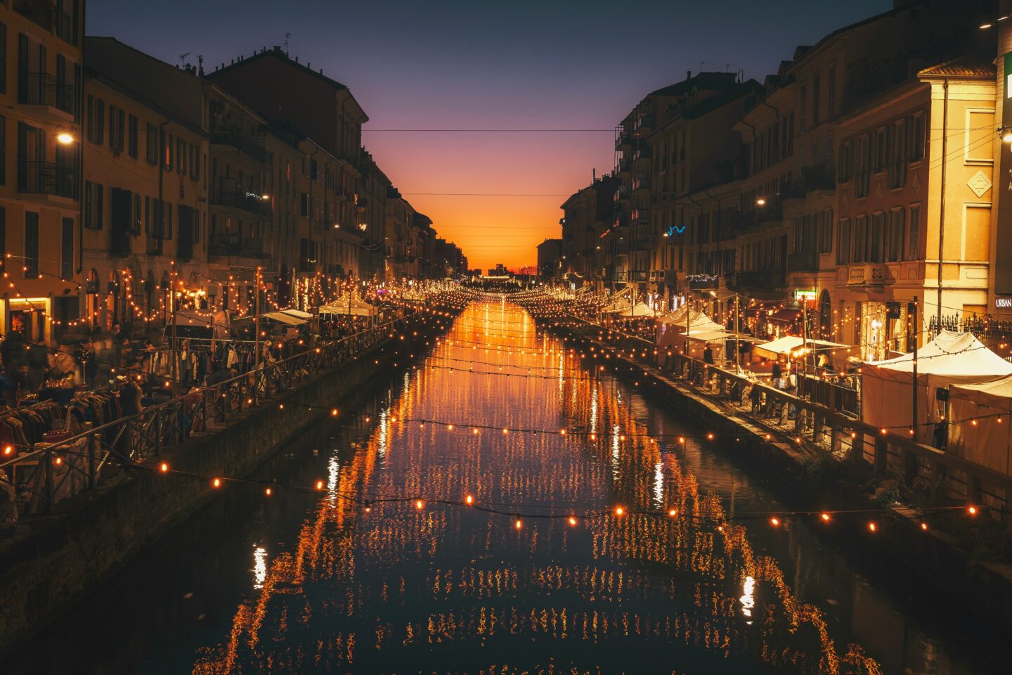 A view of Milan's Navigli Canal at sunset, beautifully decorated with festive string lights that reflect on the water, surrounded by market stalls and bustling holiday crowds along the canal's edge under a vibrant evening sky.