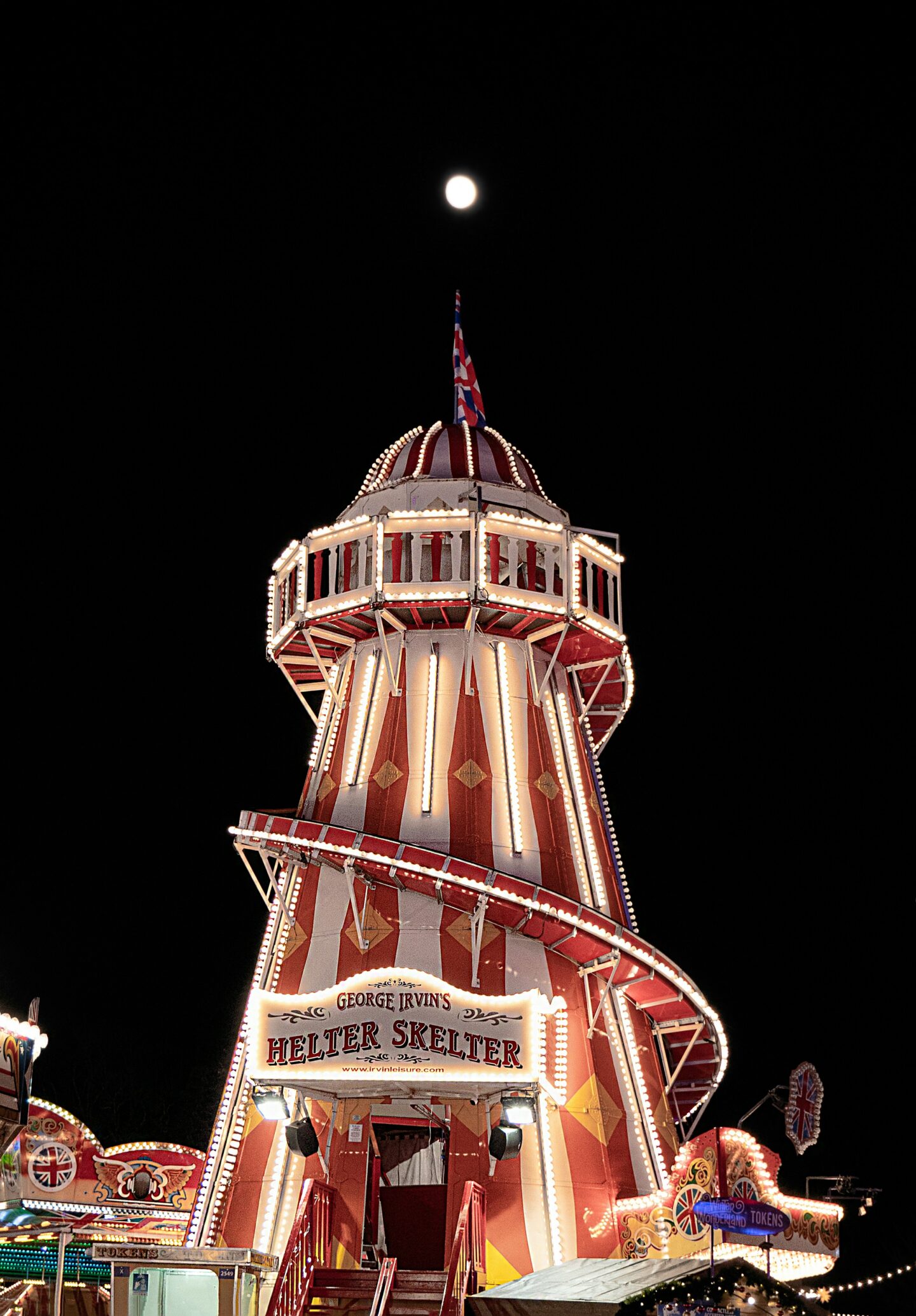 This image captures the iconic red and white Helter Skelter at Hyde Park Winter Wonderland, illuminated with bright lights against the night sky. The Union Jack flag waves at the top of the structure, with a full moon glowing above, enhancing the festive atmosphere of the event.