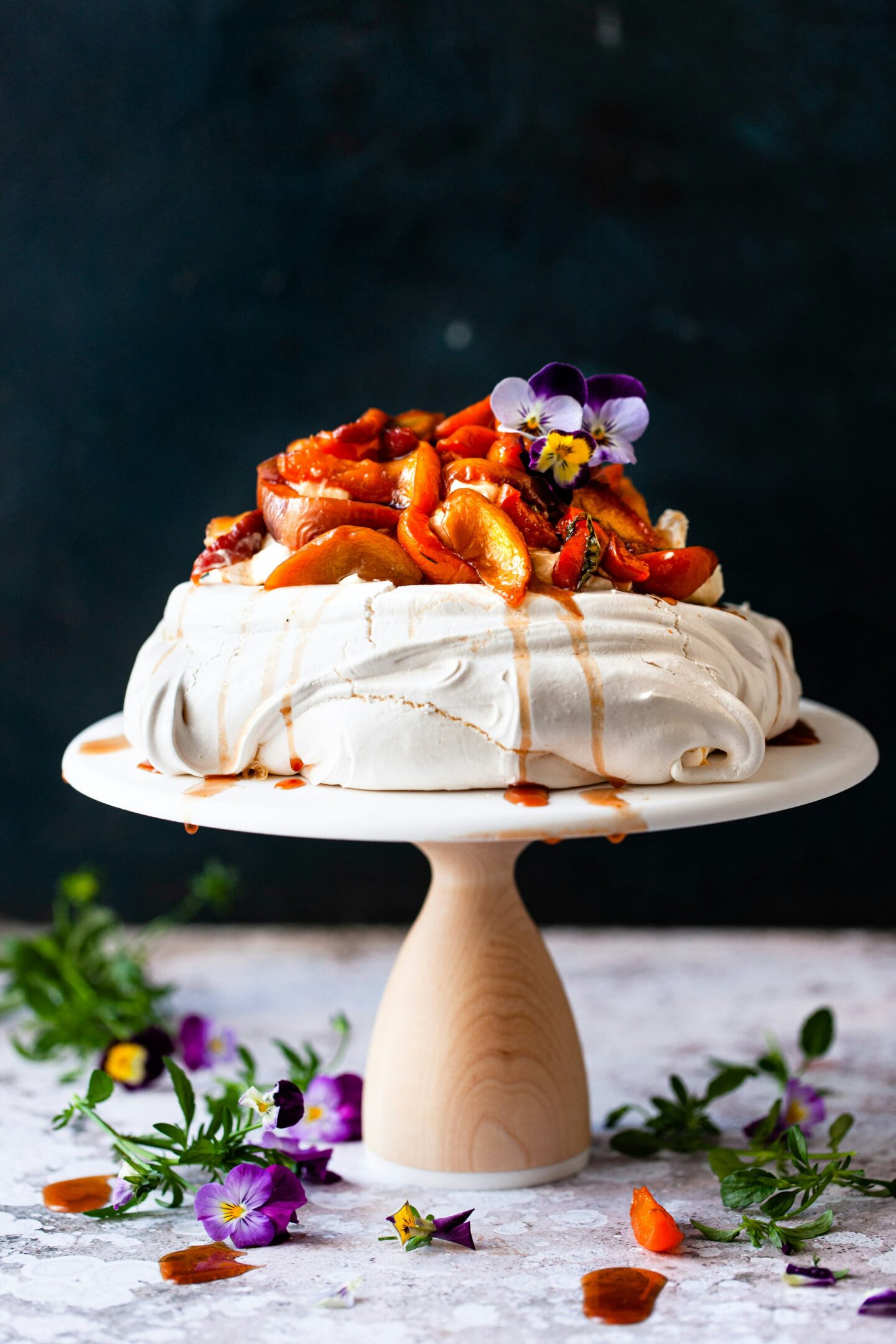 A pavlova dessert topped with peaches and garnished with edible purple flowers sits on a wooden pedestal against a dark background. The pavlova’s meringue base has delicate cracks, with sauce drizzling down, and more purple flowers scattered around the base, adding a natural, artistic touch.
