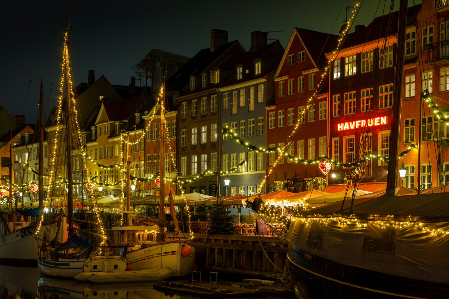 The colourful facades of Nyhavn in Copenhagen decorated with twinkling Christmas lights, reflecting off the water and surrounding boats, creating a warm holiday ambiance in the historic harbor area.