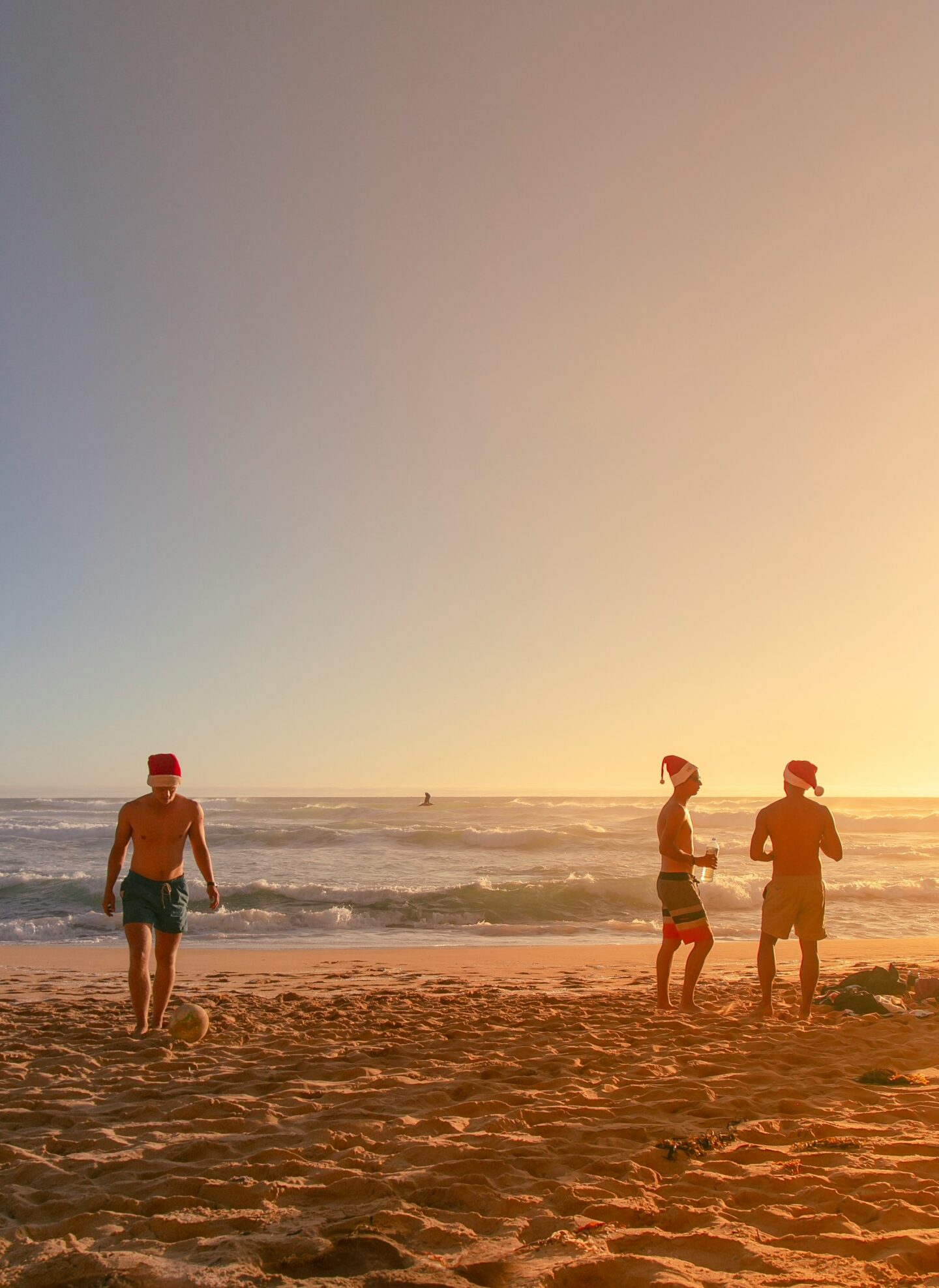Three men wearing Santa hats stand on a sandy beach during sunset, with two chatting near the ocean and one playing with a soccer ball. The warm, golden light highlights the relaxed, summery vibe, symbolizing a warm Christmas celebration typical of Australia.