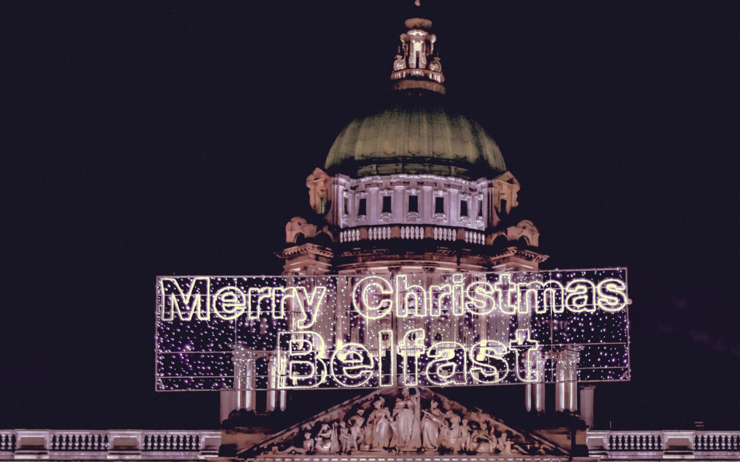 Belfast Christmas Market: Belfast City Hall lit up at night, with a bright "Merry Christmas Belfast" sign glowing in front of the iconic green dome and intricate architectural details.