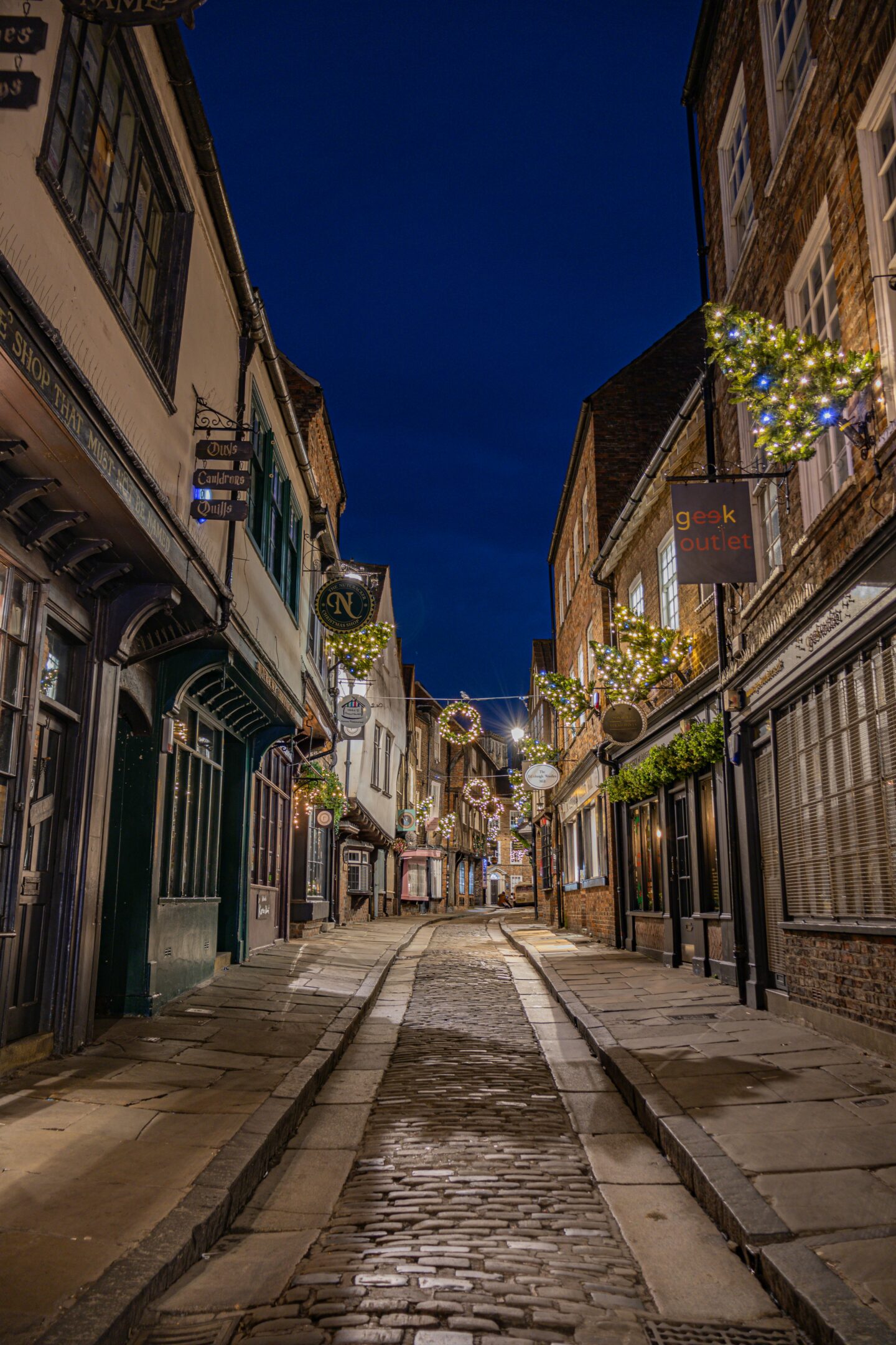 The historic Shambles in York, a cobblestone street lined with traditional timber-framed shopfronts, beautifully decorated with Christmas wreaths and glowing fairy lights for the festive season.