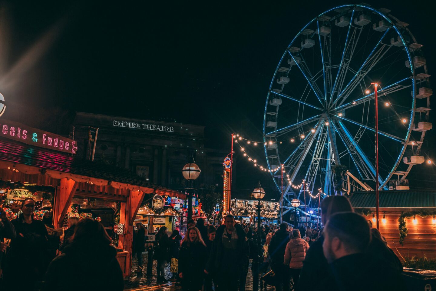 Liverpool Christmas Market: A festive scene at Liverpool Christmas Market with wooden stalls illuminated by colorful lights, a glowing Ferris wheel in the background, and the iconic Empire Theatre building lit up at night.