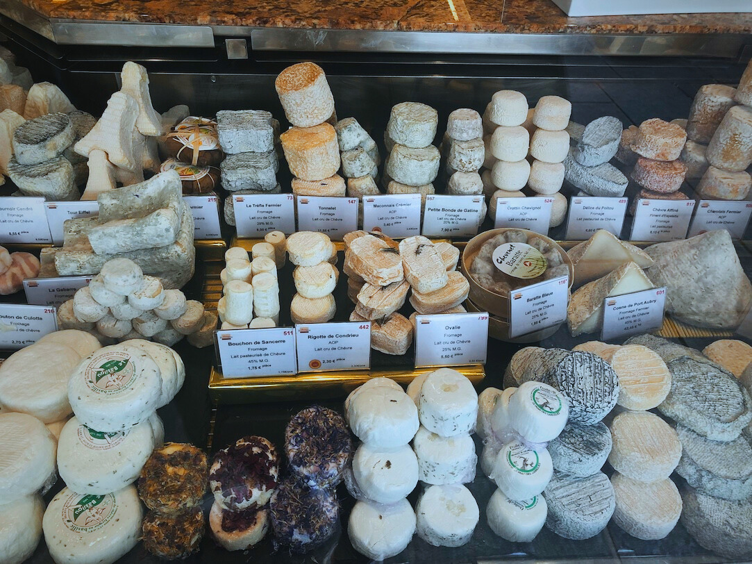 The first image showcases a beautiful cheese display at Le Marché Couvert Beauvau in Paris, France, featuring a diverse selection of artisanal French goat cheeses. The glass case is filled with various shapes, textures, and aging levels of chèvre, including soft, bloomy-rind cheeses, aged logs, and herb-crusted rounds. Small signs label each type, indicating origin and price, while the carefully stacked arrangement highlights the craftsmanship behind France’s rich cheese-making tradition.