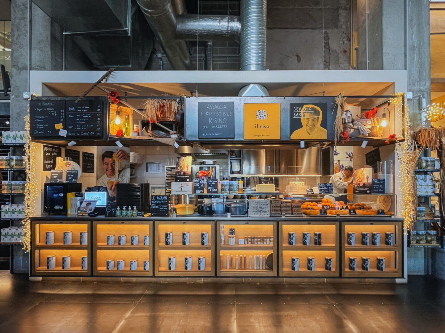 This image captures Il Riso, a charming food stall at Mercato Centrale Milano in Milan, Italy, specializing in rice-based dishes. The stand has a warm, inviting atmosphere with a modern rustic design, featuring illuminated shelves stocked with rice products and cooking essentials. A chalkboard menu lists various offerings, while signs and photos highlight Sergio Barzetti, a well-known Italian chef.