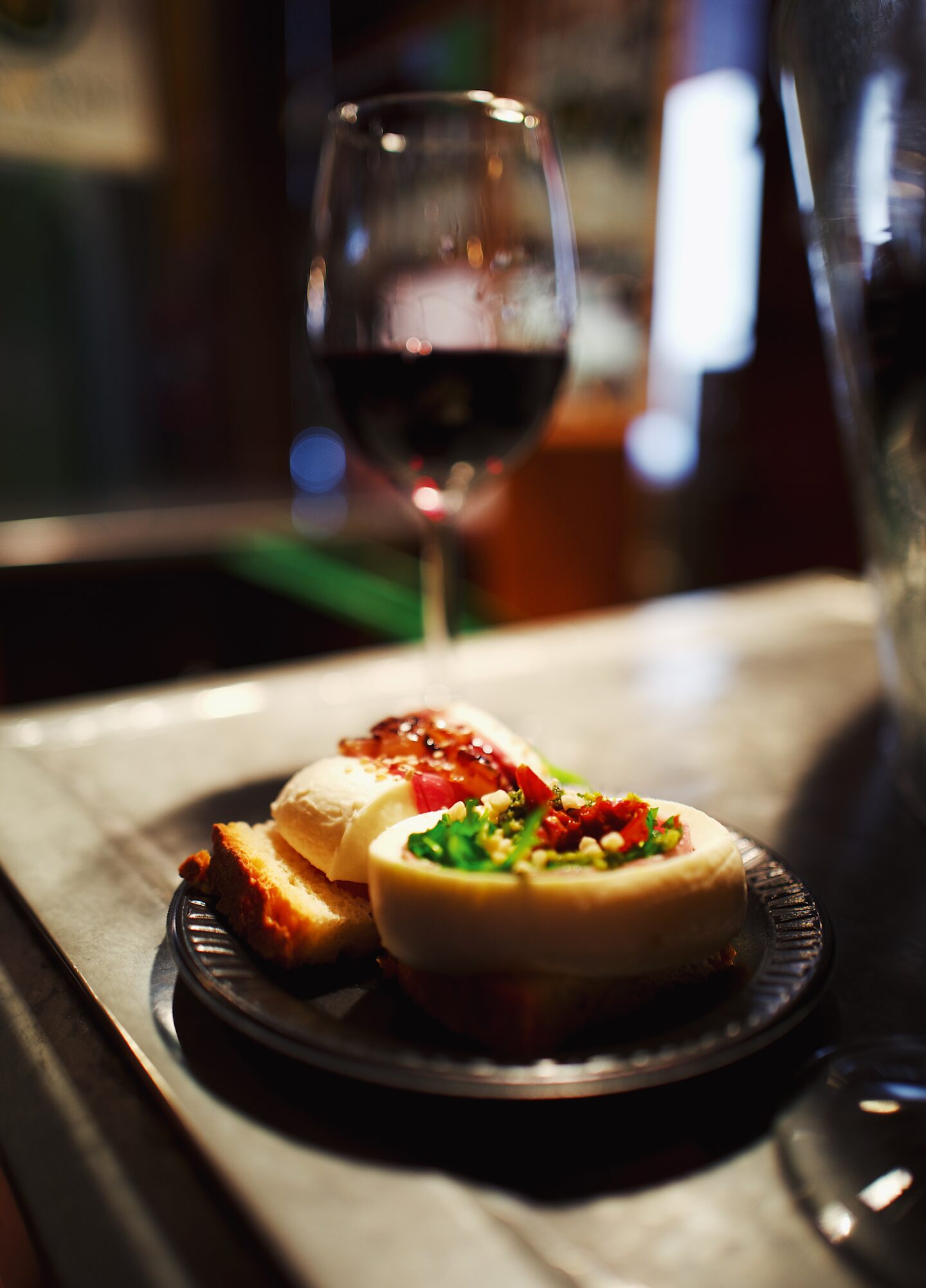 This image captures a close-up of a gourmet appetiser plate paired with a glass of red wine at Mercado de San Miguel, a famous food market in Madrid, Spain. The plate features two pieces of cheese stuffed with fresh greens, sun-dried tomatoes, and crushed nuts, along with a slice of crusty bread topped with creamy mozzarella and prosciutto. The warm lighting and blurred background create an intimate, inviting atmosphere, emphasising the rich textures and colours of the food and wine.