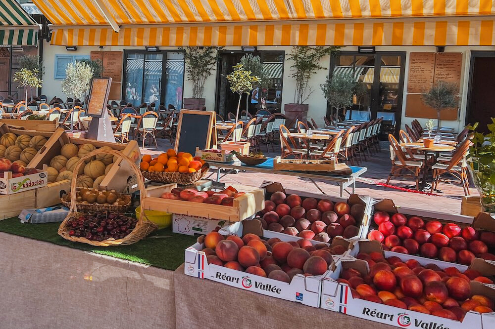 The second image captures the Cours Saleya Market in Nice, France, an open-air market famous for its fresh produce and Provençal atmosphere. The stall is filled with crates of ripe peaches, nectarines, melons, cherries, and citrus fruits, beautifully arranged under the striped yellow-and-white awning. In the background, a charming outdoor café with woven bistro chairs and handwritten menu boards invites visitors to sit and enjoy the Mediterranean ambiance. The bright colours and warm sunlight reflect the vibrant energy of this iconic French Riviera market.