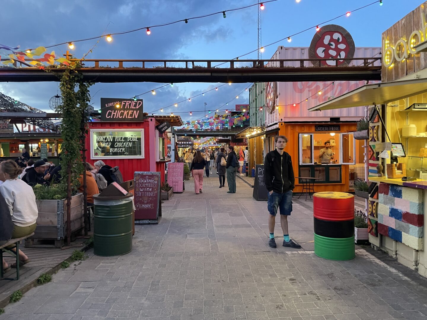 This image captures an evening scene at Reffen, a popular street food market in Copenhagen, Denmark. The market is lively with people sitting at communal tables and walking along a pathway lined with colourful food stalls made from repurposed shipping containers. String lights and decorations create a festive atmosphere, while various signs advertise international cuisine, including Korean fried chicken and cocktails. A young man in casual attire stands near a red and green barrel, adding a human element to the vibrant setting.