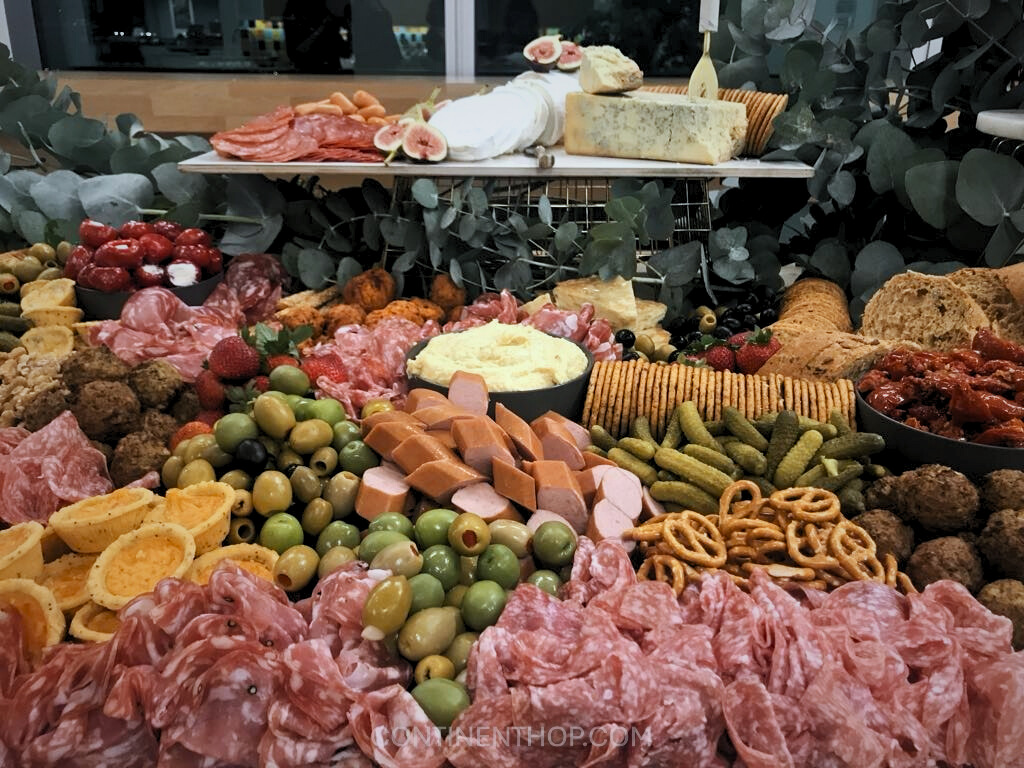 A stunning charcuterie spread at the Rialto Market in Venice, Italy. The table is overflowing with Italian delicacies, including an assortment of cured meats like salami and prosciutto, various cheeses such as blue cheese and brie, and an array of antipasti including olives, pickles, sun-dried tomatoes, and stuffed cherry peppers. Crackers, pretzels, and fresh bread slices complement the selection, while figs and strawberries add a touch of sweetness.