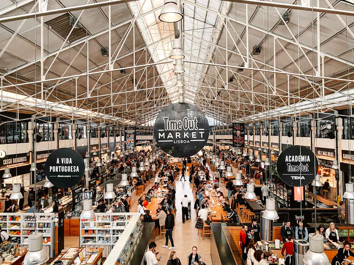 The image showcases the bustling Time Out Market in Lisbon, Portugal, a vibrant food hall housed in a spacious industrial-style building with a high ceiling and exposed metal framework. Large black circular signs hang from the ceiling, displaying names of various vendors and sections, including "A Vida Portuguesa" and "Academia Time Out." The market is filled with people enjoying diverse culinary offerings at communal tables, while food stalls line both sides, serving a mix of traditional Portuguese dishes and international cuisine. The bright and lively atmosphere reflects the market's reputation as a top gastronomic destination.