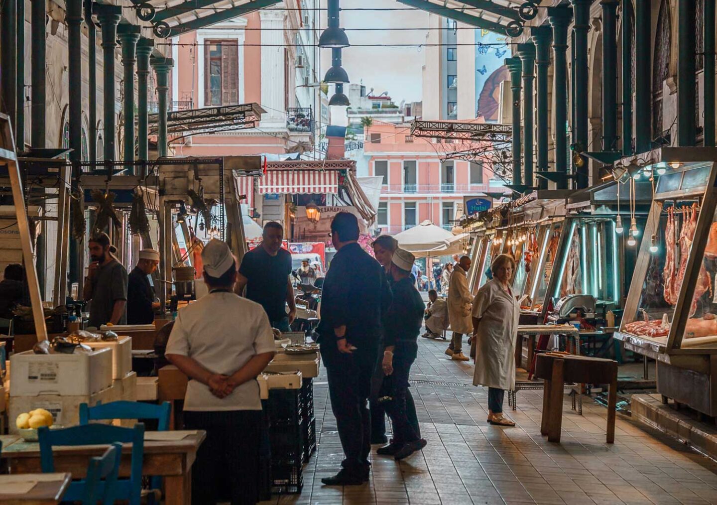 This image captures the lively Varvakios Market in Athens, Greece, a historic central market known for its fresh meat, seafood, and produce. The covered walkway is lined with butcher stalls displaying cuts of meat under bright lights, while vendors in white coats engage with customers. In the foreground, a chef stands with hands behind their back, observing the market’s activity. The market’s rustic charm, with its iron columns and hanging light bulbs, contrasts with the bright, colourful buildings visible in the background. The scene reflects the bustling, authentic atmosphere of this iconic Athenian food market.