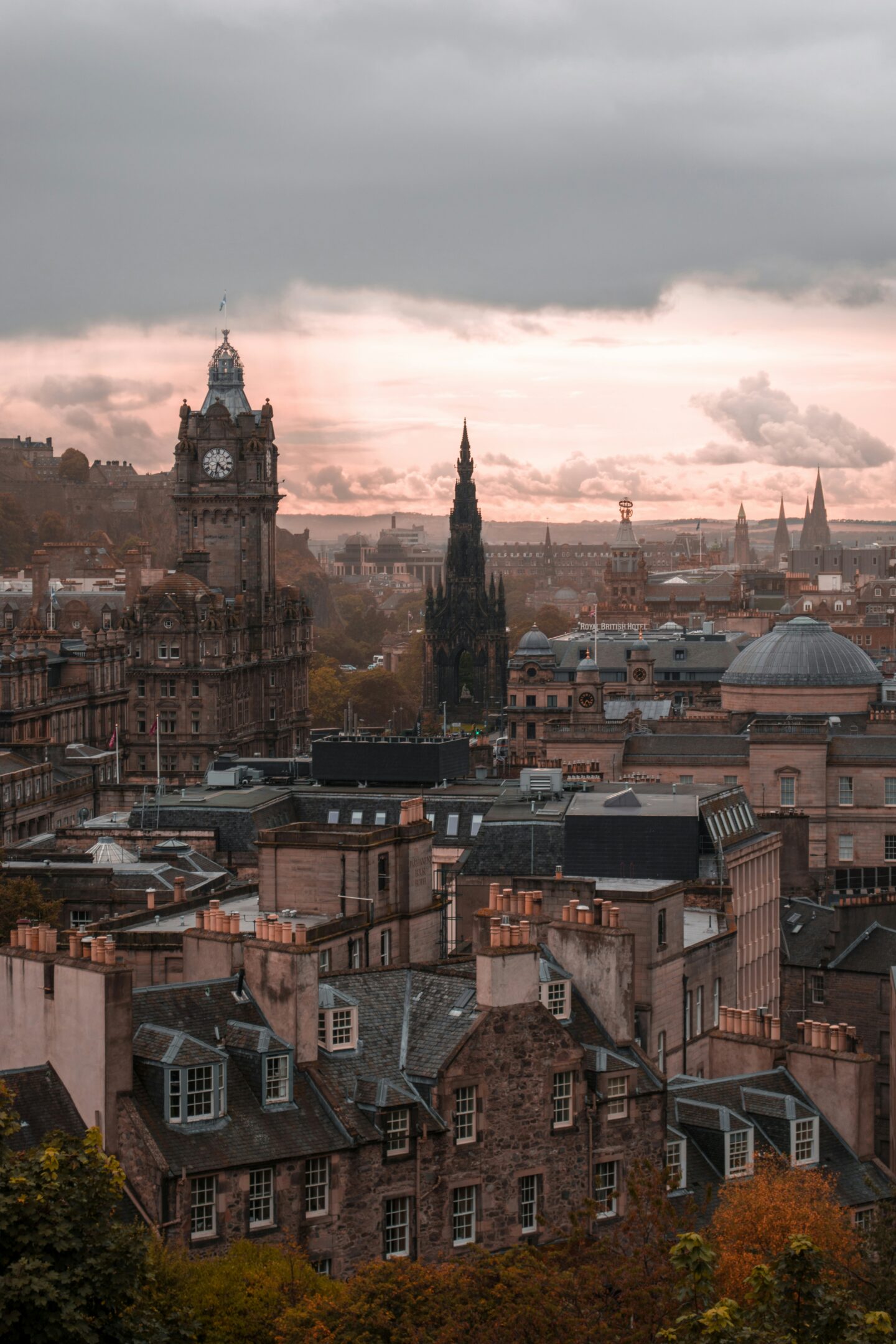 A dramatic view of Edinburgh’s cityscape at sunset, featuring the iconic Balmoral Hotel clock tower and the Gothic spire of the Scott Monument. The historic stone buildings and rooftops are bathed in soft, dusky light, with a backdrop of rolling hills and a moody sky. The scene captures the timeless charm and architectural beauty of Scotland’s capital city.