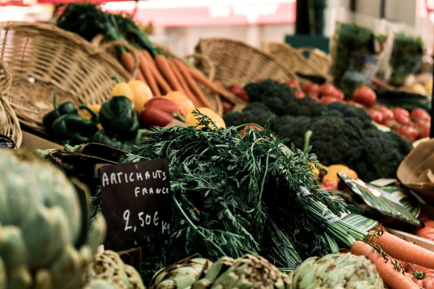 This image showcases a vibrant French farmers' market stall, featuring an abundance of fresh vegetables displayed in woven baskets. In focus is a handwritten chalkboard sign reading "Artichauts France – 2,50€/kg," surrounded by lush green carrots, artichokes, bell peppers, broccoli, and ripe tomatoes. The earthy tones and natural textures create a rustic and inviting atmosphere, reflecting the charm of local open-air markets in France. The blurred background hints at more produce and the lively market setting, where shoppers can find high-quality, seasonal ingredients.