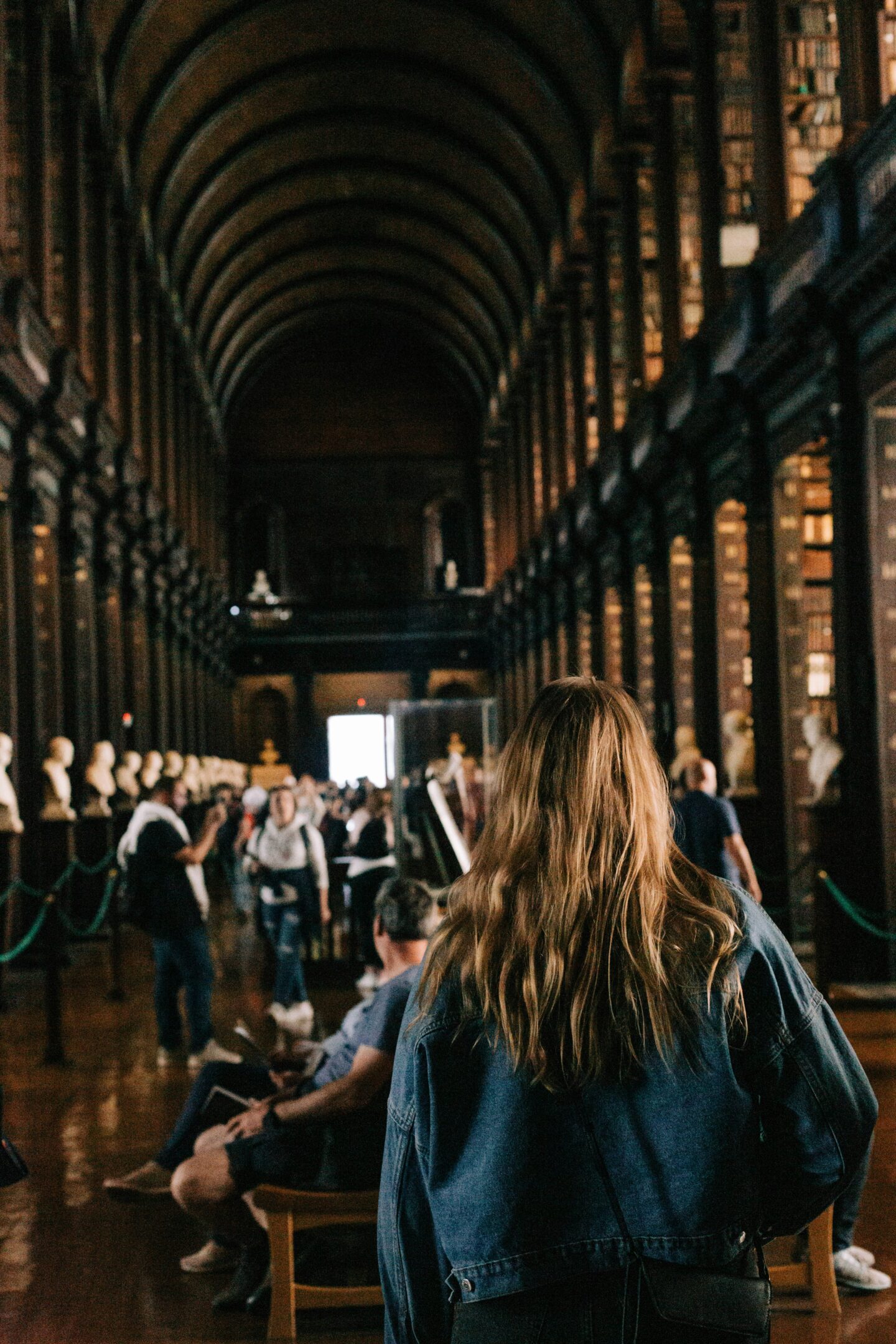 A woman with long, wavy blonde hair in a denim jacket walks through the Long Room of the Trinity College Library in Dublin, home to the Book of Kells. The historic hall features towering bookshelves, marble busts, and a vaulted ceiling, with visitors admiring the iconic library’s rich history. The scene captures the quiet reverence and scholarly atmosphere of one of Ireland's most famous cultural landmarks.