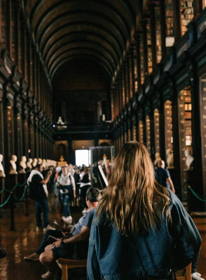 A woman with long, wavy blonde hair in a denim jacket walks through the Long Room of the Trinity College Library in Dublin, home to the Book of Kells. The historic hall features towering bookshelves, marble busts, and a vaulted ceiling, with visitors admiring the iconic library’s rich history. The scene captures the quiet reverence and scholarly atmosphere of one of Ireland's most famous cultural landmarks.