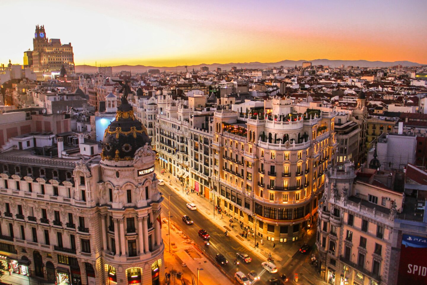 A stunning aerial view of Madrid’s Gran Vía at sunset, showcasing the iconic Metropolis building with its ornate dome and golden details. The streets below are illuminated by warm lights as traffic and pedestrians move through the bustling city. The vibrant sky transitions from orange to purple hues, with distant mountains silhouetted on the horizon.