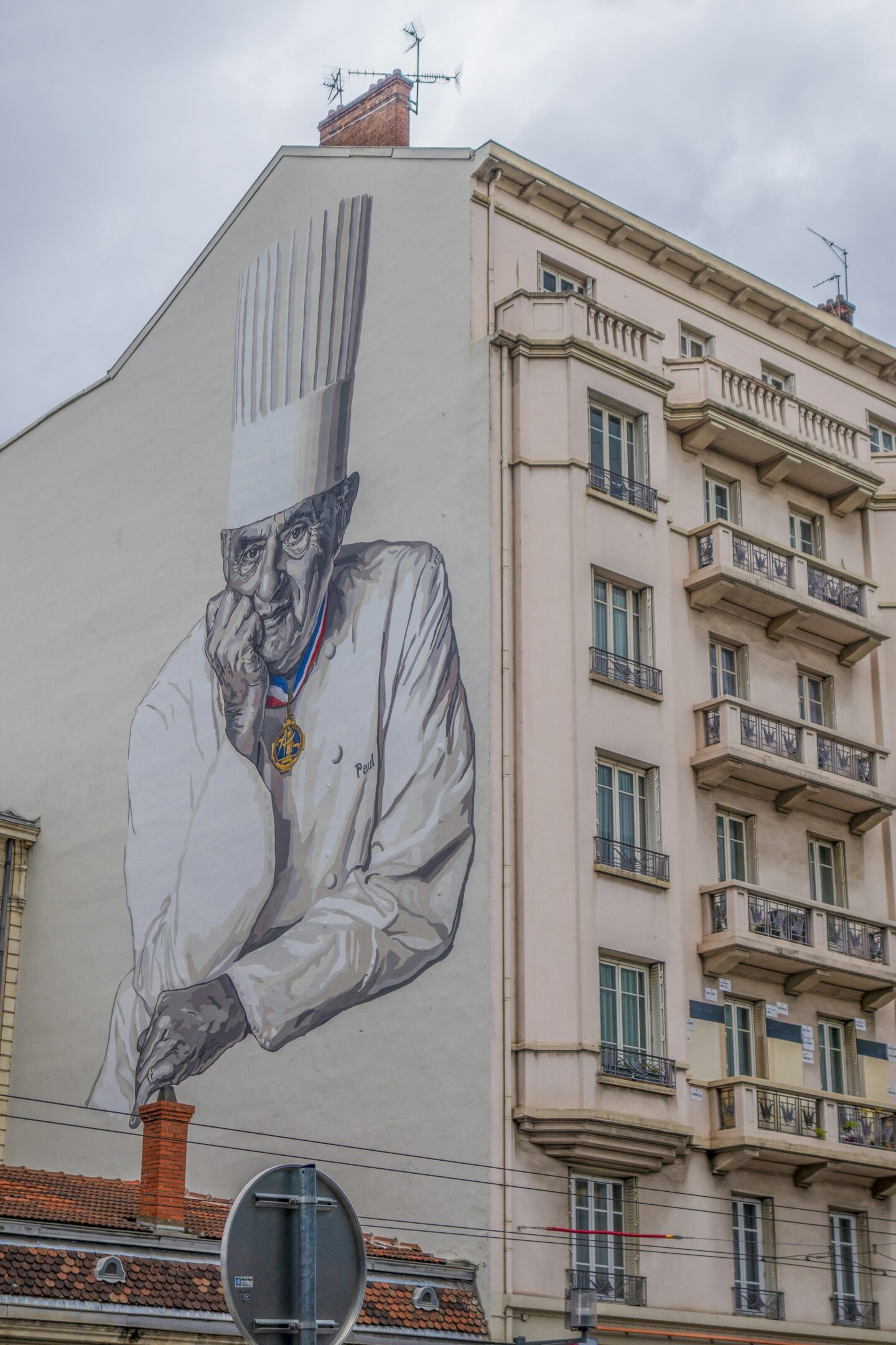 This image showcases a large mural of Paul Bocuse, the renowned French chef, painted on the side of a building in Lyon, France. The monochromatic artwork depicts him in his signature chef’s attire, including a tall white toque, with a thoughtful expression and his hand resting on his chin. The mural, created by Cité Création and students of Ecocité in 2015, is located in the 3rd arrondissement, near the Halles Paul Bocuse, paying tribute to his culinary legacy.