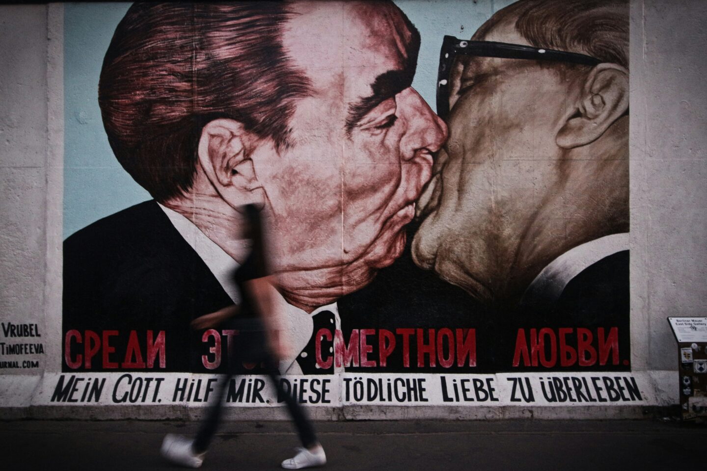 A famous mural from the Berlin Wall depicting a symbolic kiss between two male political leaders, with bold text in Russian and German that translates to “My God, help me survive this deadly love.” A blurred figure walks past the mural, adding motion to the scene against the historical artwork.