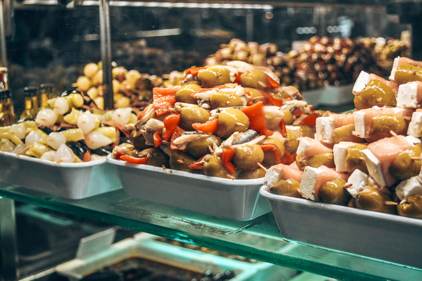 This image captures a vibrant display of pintxos and tapas at Mercado de la Paz in Madrid, Spain, a well-known traditional food market. The glass case is filled with skewered green olives, anchovies, roasted red peppers, and cheese wrapped in prosciutto, all glistening with olive oil. The selection showcases the Basque and Spanish tradition of bite-sized gourmet snacks, served fresh and ready to enjoy. The rich colours and textures highlight the high-quality ingredients that make Madrid’s food markets a paradise for food lovers.