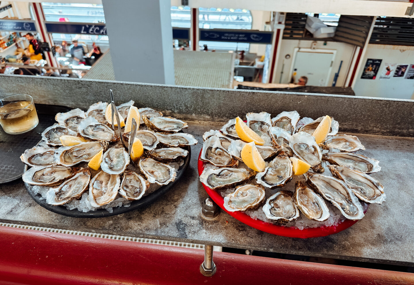 This image showcases two beautifully arranged platters of fresh oysters on ice, garnished with lemon wedges, served at Les Halles de Biarritz, a renowned food market in Biarritz, France. The oysters are presented on black and red trays, accompanied by small forks for easy eating. In the background, the market atmosphere is visible, with seafood stalls and people enjoying the vibrant culinary scene. A glass of white wine sits beside the oysters, highlighting the perfect pairing of seafood and wine in this classic Basque coastal experience.