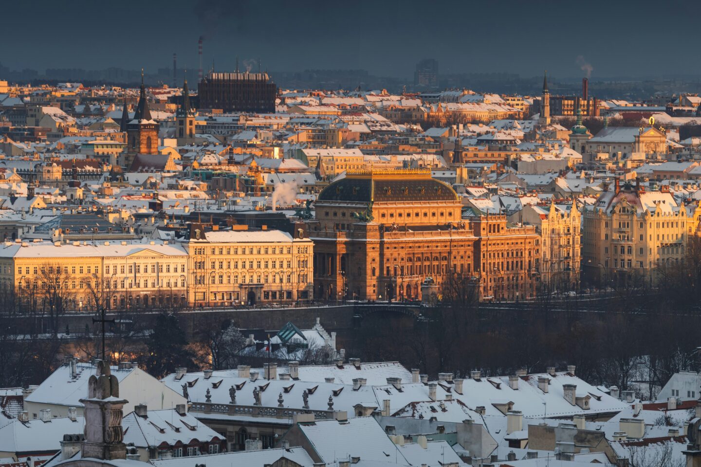 A panoramic winter view of Prague, with the historic National Theatre standing out as the sunlight casts a golden glow on its façade. The surrounding rooftops are covered in snow, creating a striking contrast between the warm light and the cold, wintry scene. The cityscape features a blend of Gothic and Baroque architecture, with church spires and historic buildings adding to Prague’s timeless charm.
