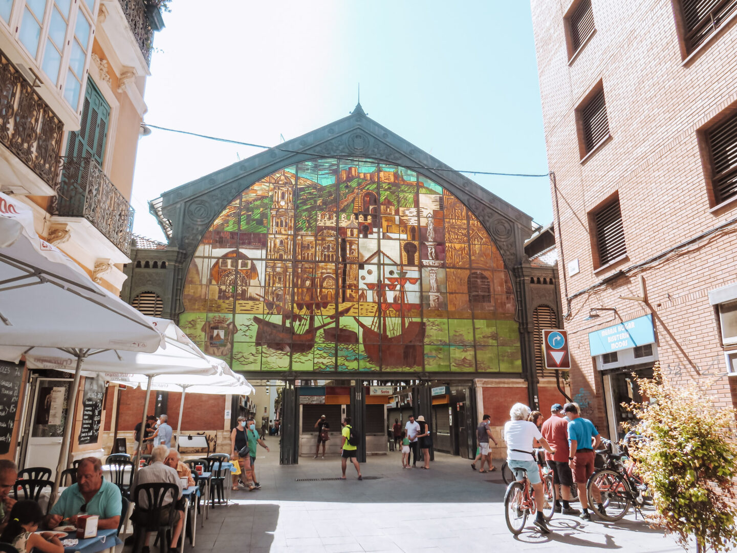 This image captures the Atarazanas Market (Mercado Central de Atarazanas) in Málaga, Spain, a historic and bustling food market. The focal point is the beautiful stained-glass window, which showcases a vibrant depiction of Málaga’s landmarks, ships, and coastal scenery. The market's iron-and-glass architecture reflects its 19th-century industrial design, blending history with modern commerce. Outside, people are seen enjoying meals at sidewalk cafés under white umbrellas, while others walk or ride bicycles. The lively yet relaxed atmosphere embodies the essence of Southern Spain’s market culture, where locals and visitors alike gather to enjoy fresh produce, seafood, and tapas.