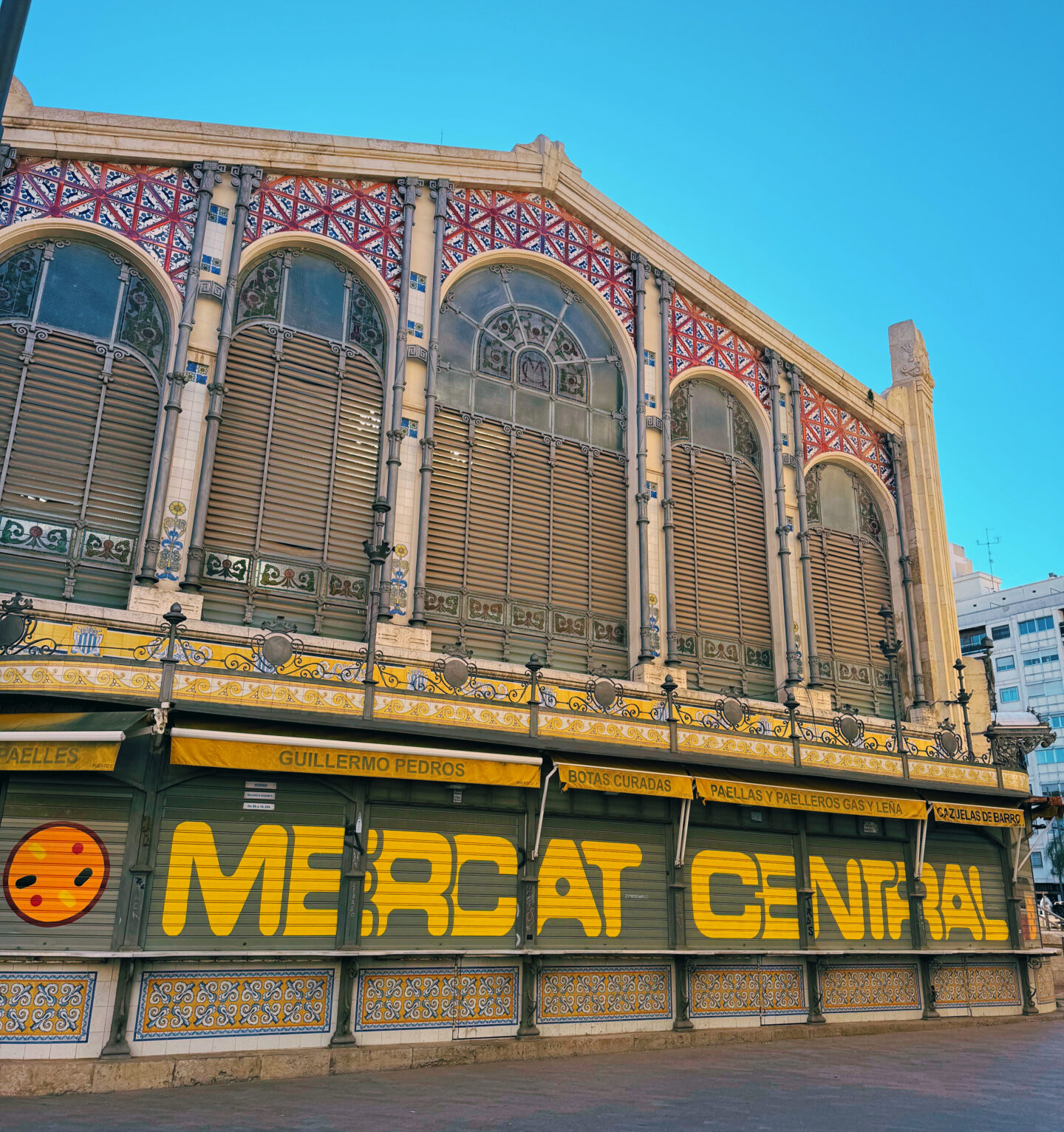 The image showcases the Mercado Central (Central Market) in Valencia, Spain, one of the largest and most beautiful indoor markets in Europe. The stunning façade features ornate stained-glass windows, colourful ceramic tiles, and intricate ironwork, reflecting the city's rich Modernist architectural heritage. The bold yellow lettering on the shutters contrasts with the historic detailing, creating a mix of old and new. This market is a culinary hub, offering fresh produce, seafood, meats, and local specialties, making it a must-visit destination for food lovers in Valencia.
