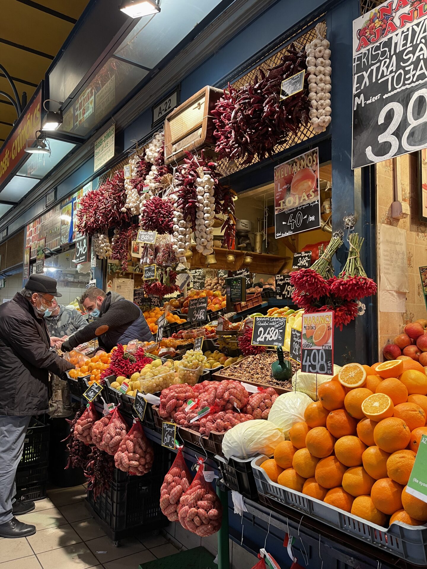 A bustling scene inside Budapest’s Great Market Hall, featuring rows of stalls filled with fresh produce, meats, cheeses, and Hungarian specialties. Shoppers browse under the grand, high-ceilinged structure with intricate ironwork and large windows that let in natural light. Vendors display paprika, sausages, and traditional goods, creating a vibrant atmosphere in this historic market.