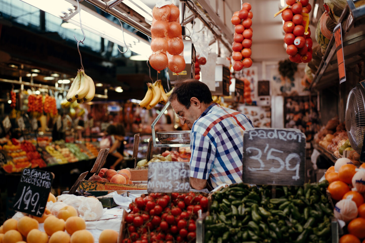 A vibrant fruit and vegetable stall at La Boqueria Market in Barcelona, with a vendor in a checkered shirt arranging fresh produce. The stall is filled with ripe peaches, cherry tomatoes, green padrón peppers, and hanging bananas, while bunches of tomatoes and oranges dangle from above. Handwritten chalkboard signs display prices, adding to the authentic market atmosphere.